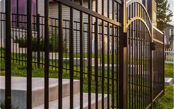 A black ornamental metal fence with an arched gate next to concrete steps and a lawn in a residential area.