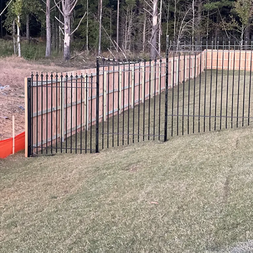 A view of a newly installed backyard perimeter fence on a sloped grassy area, featuring a combination of natural wood privacy panels and black ornamental aluminum fencing with decorative spear-top finials along the front section.