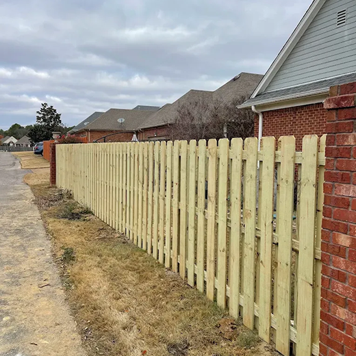 A view of a newly installed semi-privacy picket fence featuring vertical pressure-treated wooden slats with dog-ear tops, spanning along a residential property line between brick pillars and a paved walkway under an overcast sky.
