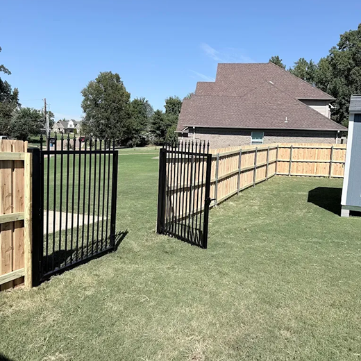 A view of a newly installed backyard enclosure featuring a combination of a natural wood privacy fence and a black ornamental metal double gate with decorative finials, bordering a large green lawn behind a residential home.